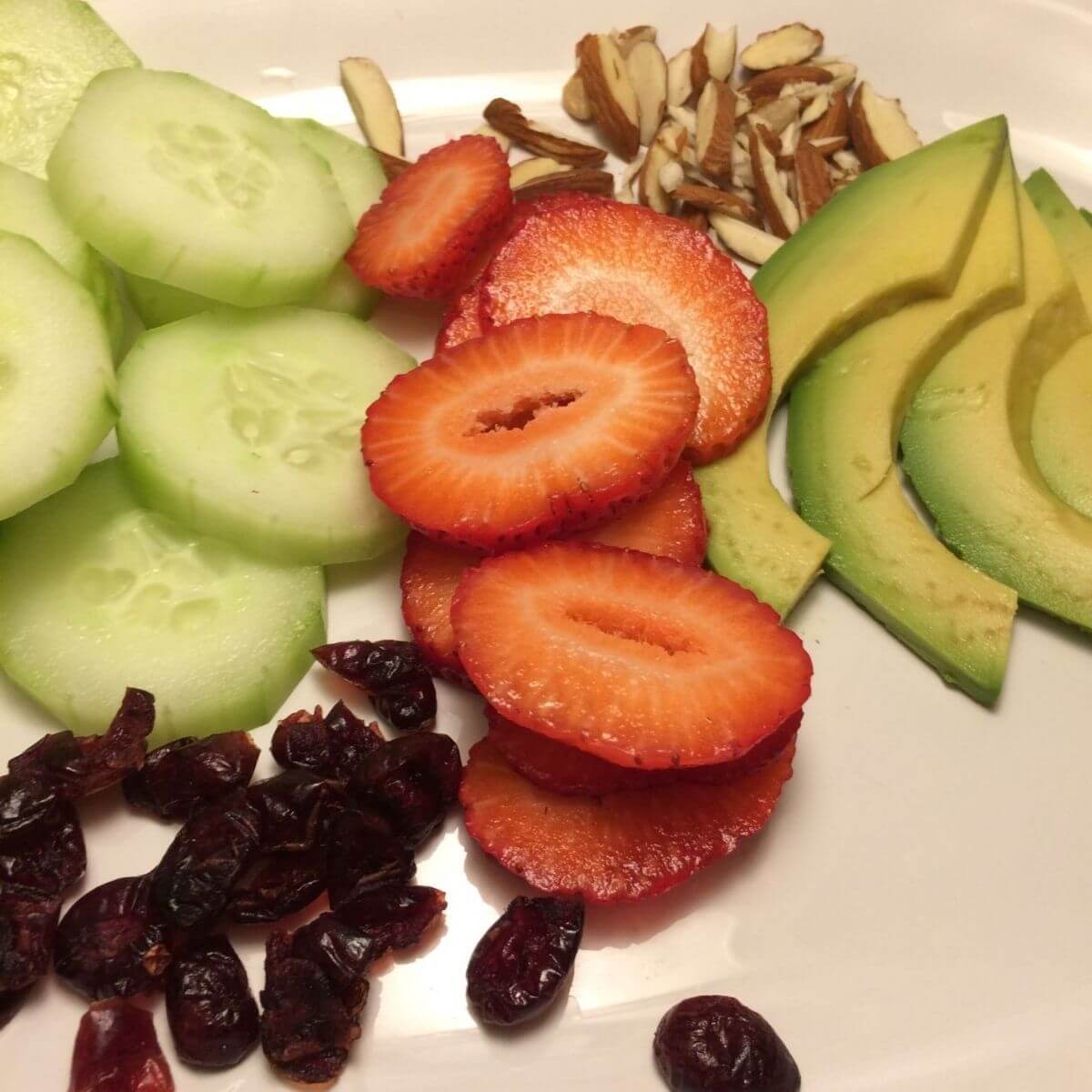 sliced cucumbers, strawberries, avocados, almonds, and whole dried craisins on a white plate