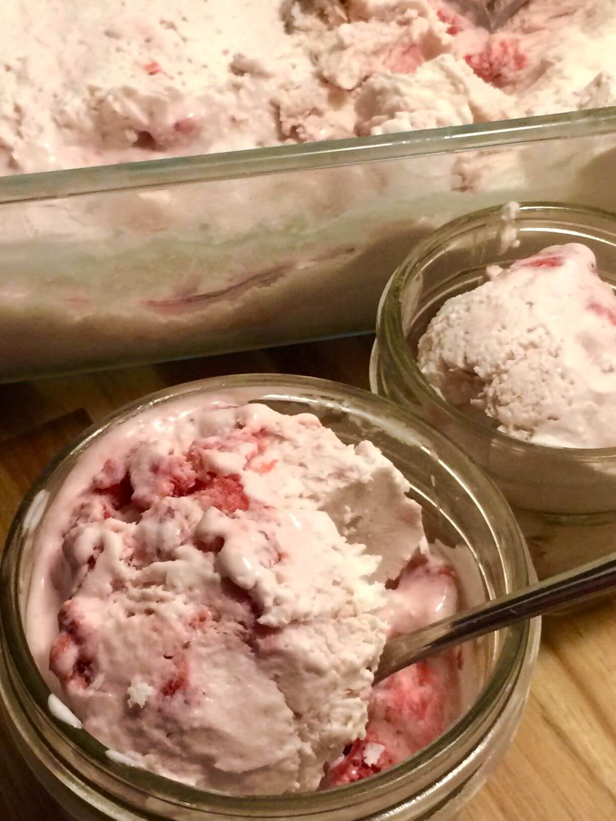 Two 8 ounce mason jars with scoops of strawberry ice cream, 1 with a spoon, with 11 cup glass pyrex with remaining ice cream in the background.