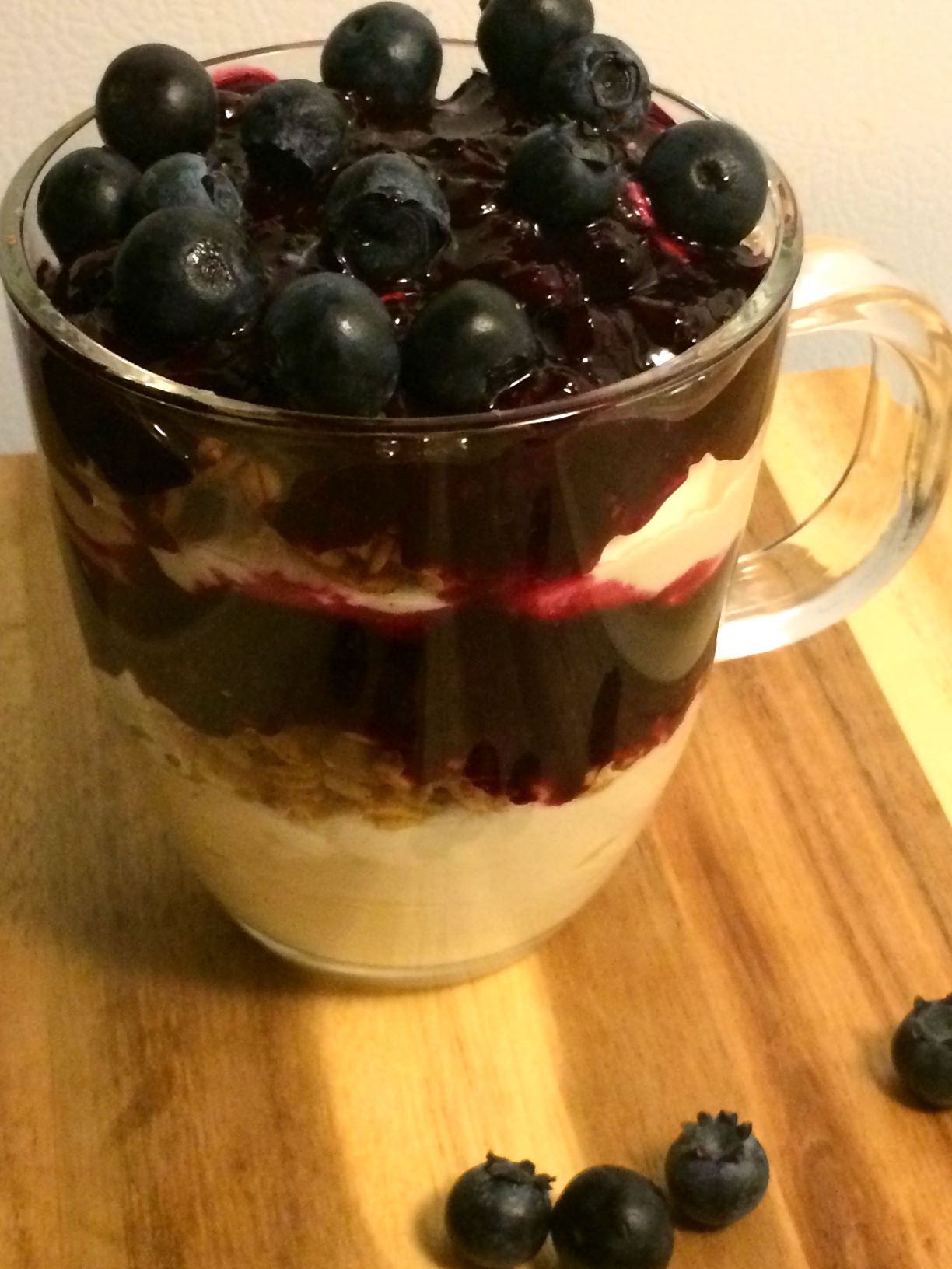 glass mug with layered mascarpone cream, blueberry pie filling, and granola all on a wood cutting board with fresh blueberries scattered on board.