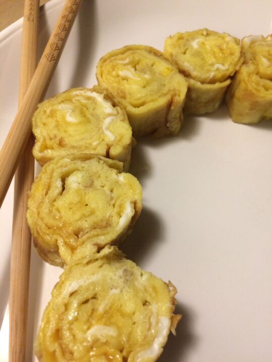 tamago rolled up into 6 rounds and slightly arched like a rainbow with chopstick to the left, all on a white plate.