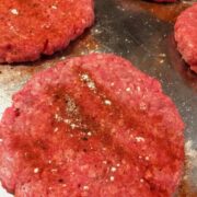 raw hamburger patty rounds with seasoning on top on stainless steel baking sheet