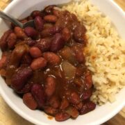 cowboy baked beans in a white bowl with brown rice with metal spoon in left side of bowl all on wood board