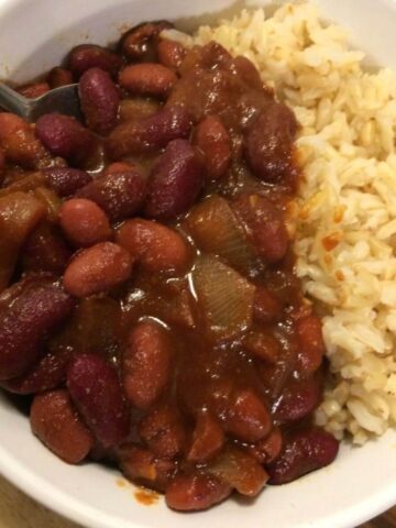 cowboy baked beans in a white bowl with brown rice with metal spoon in left side of bowl all on wood board