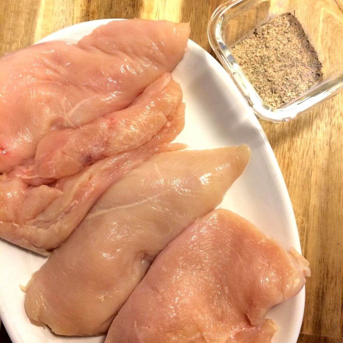 chicken breasts filleted on white oval plate with seasonings on the side in a glass square jar all on a wooden cutting board