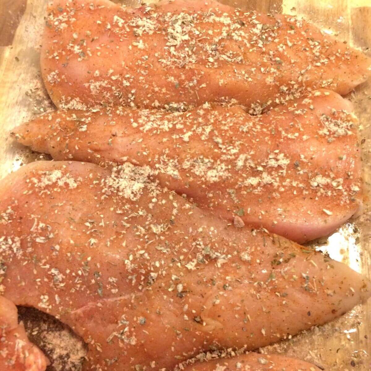 chicken breasts filleted lined side by side in glass cake pan with seasonings sprinkled on top
