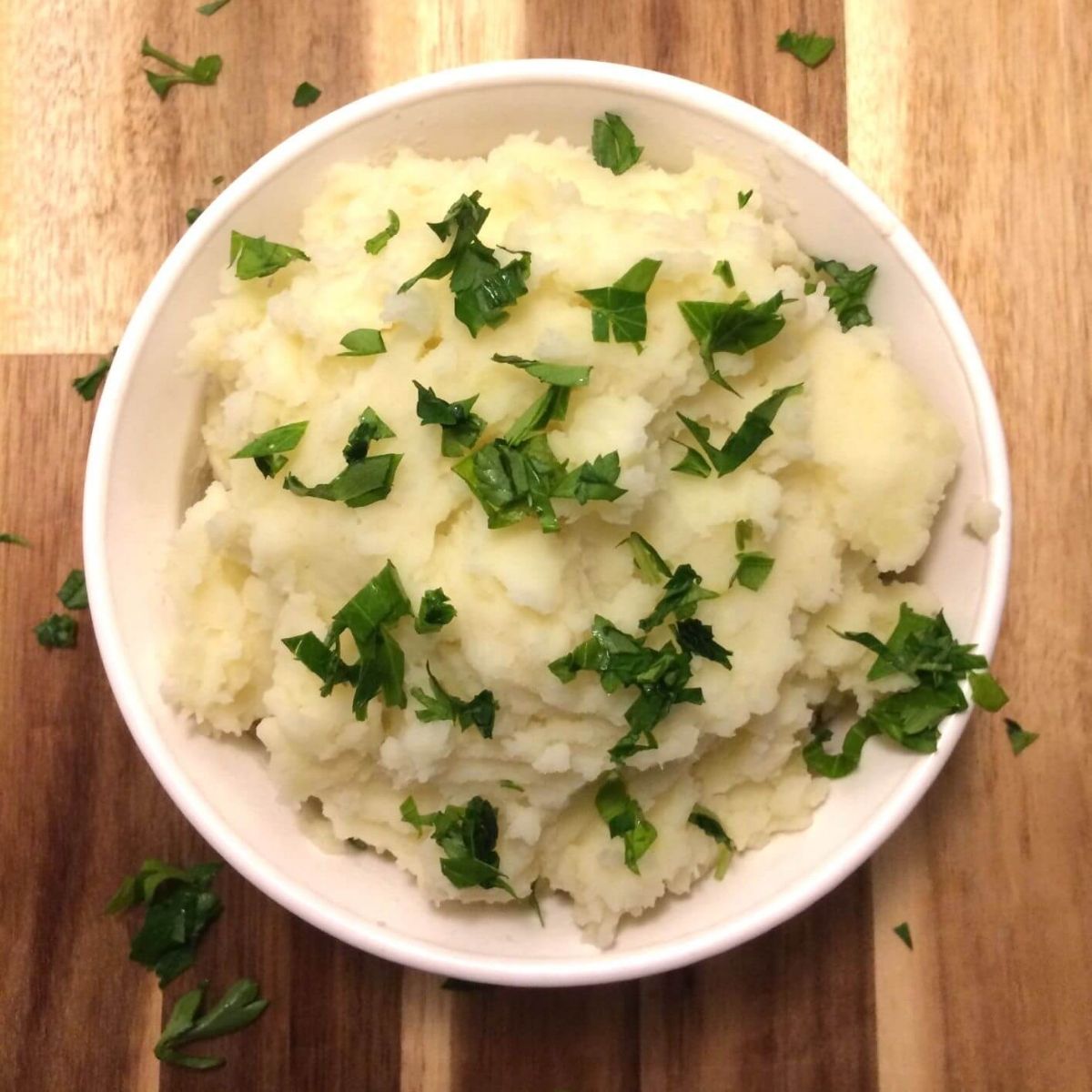 mashed potatoes garnished with fresh parsley in a white round bowl on a wooden cutting board