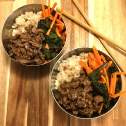 Two mongolian beef bowls with ingredients separated in stainless steel bowls with chopsticks on the side all on a wooden cutting board