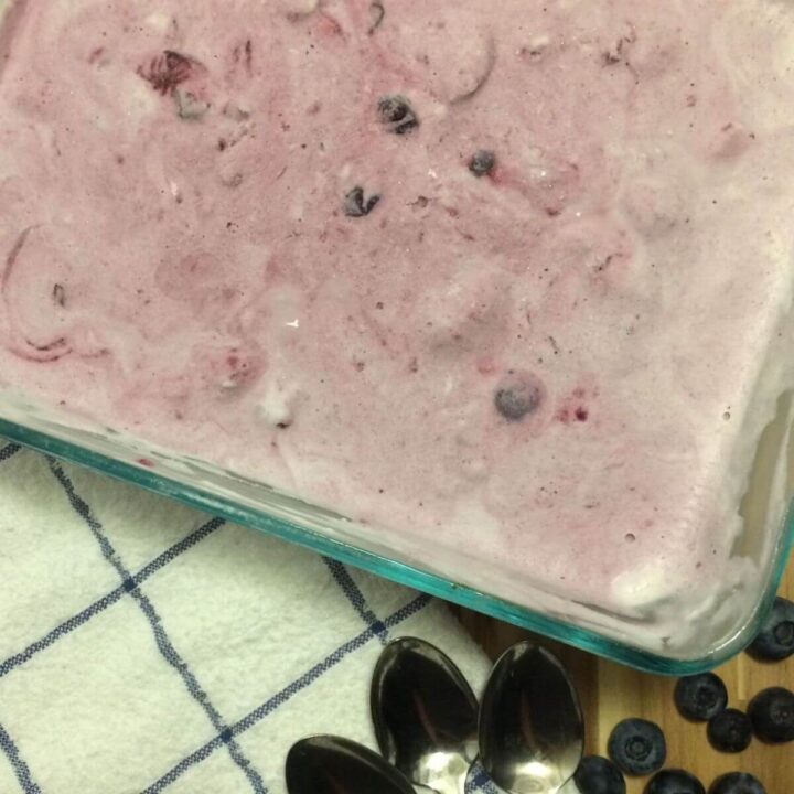 Freezing ice cream mixture in glass rectangle Pyrex with blue and white checkered tower in lower left corner with 3 silver spoons and scattered blueberries in lower right corner.