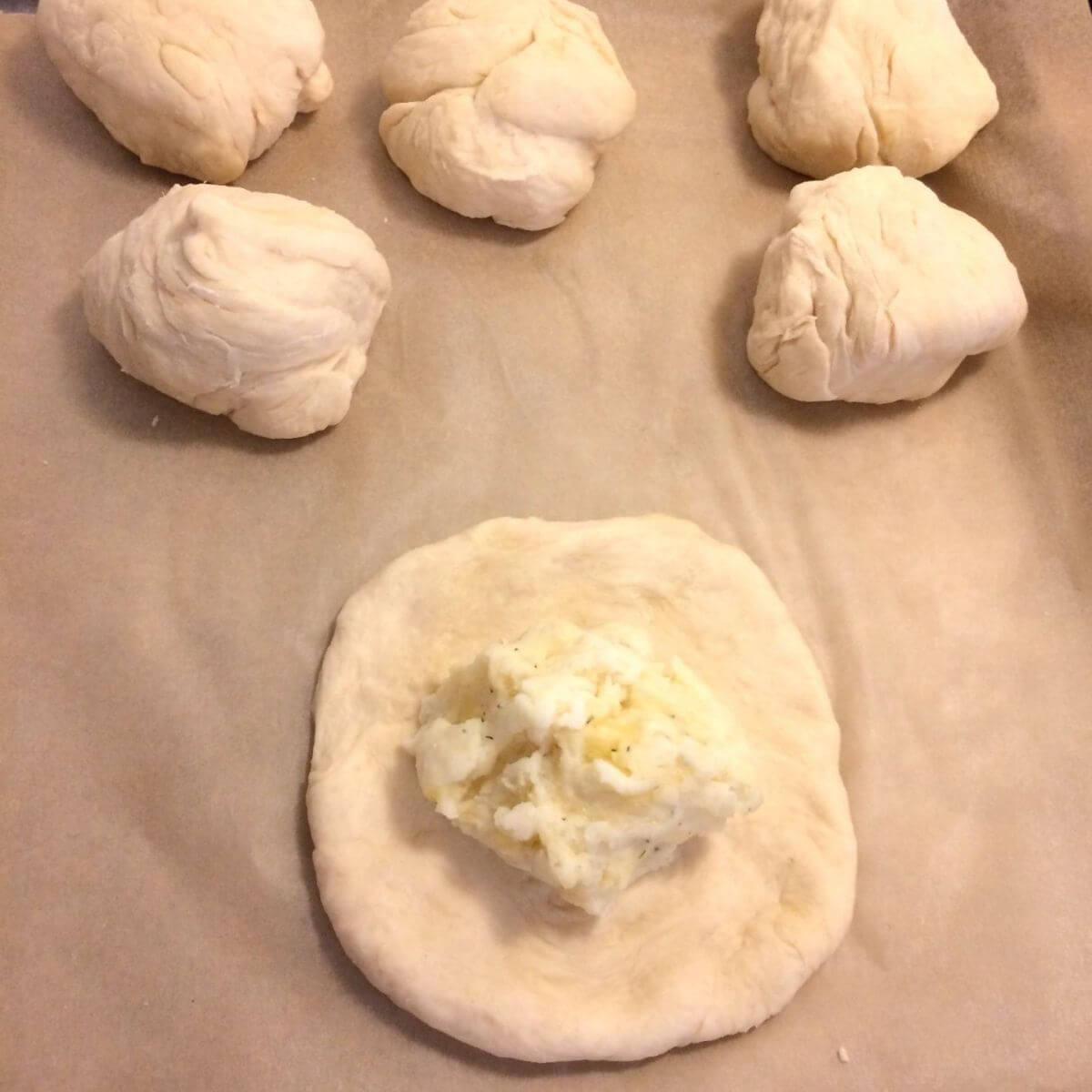 5 dough balls in the background on parchment paper with 1 dough ball stretched out into a round with filling placed in the middle.