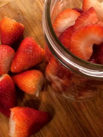 de-stemmed strawberries on a wood cutting board next to a tall mason jar with chopped up strawberries