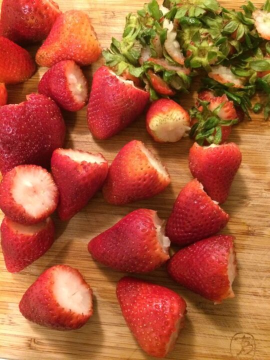 de-stemmed strawberries on a wooden cutting board with cut stems piled in the upper right corner