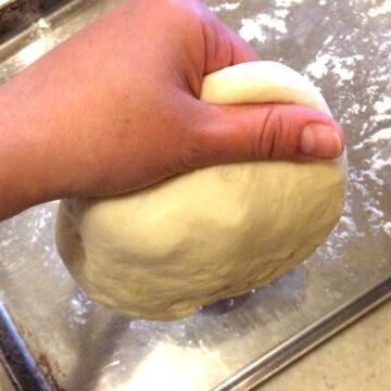 Left hand holding pizza dough with a lateral pinch in the air above baking sheet with flour sprinkled all over.