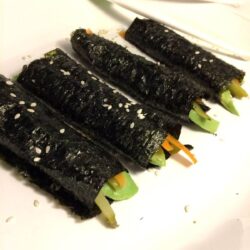 4 rolls with seaweed paper rolled around vegetables lined up on a diagonal with kid's white chopsticks in background all on a white oval plate
