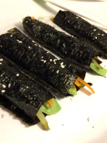 4 rolls with seaweed paper rolled around vegetables lined up on a diagonal with kid's white chopsticks in background all on a white oval plate