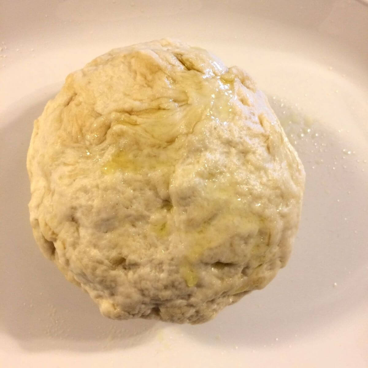 pizza dough ball with olive oil on top rolled into a ball after kneading on white plate