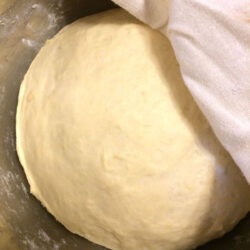 pizza dough rising in stainless steel bowl with tea towel partially covering on upper right corner