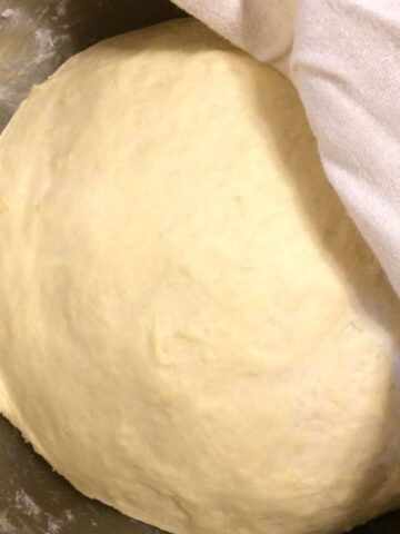 pizza dough rising in stainless steel bowl with tea towel partially covering on upper right corner