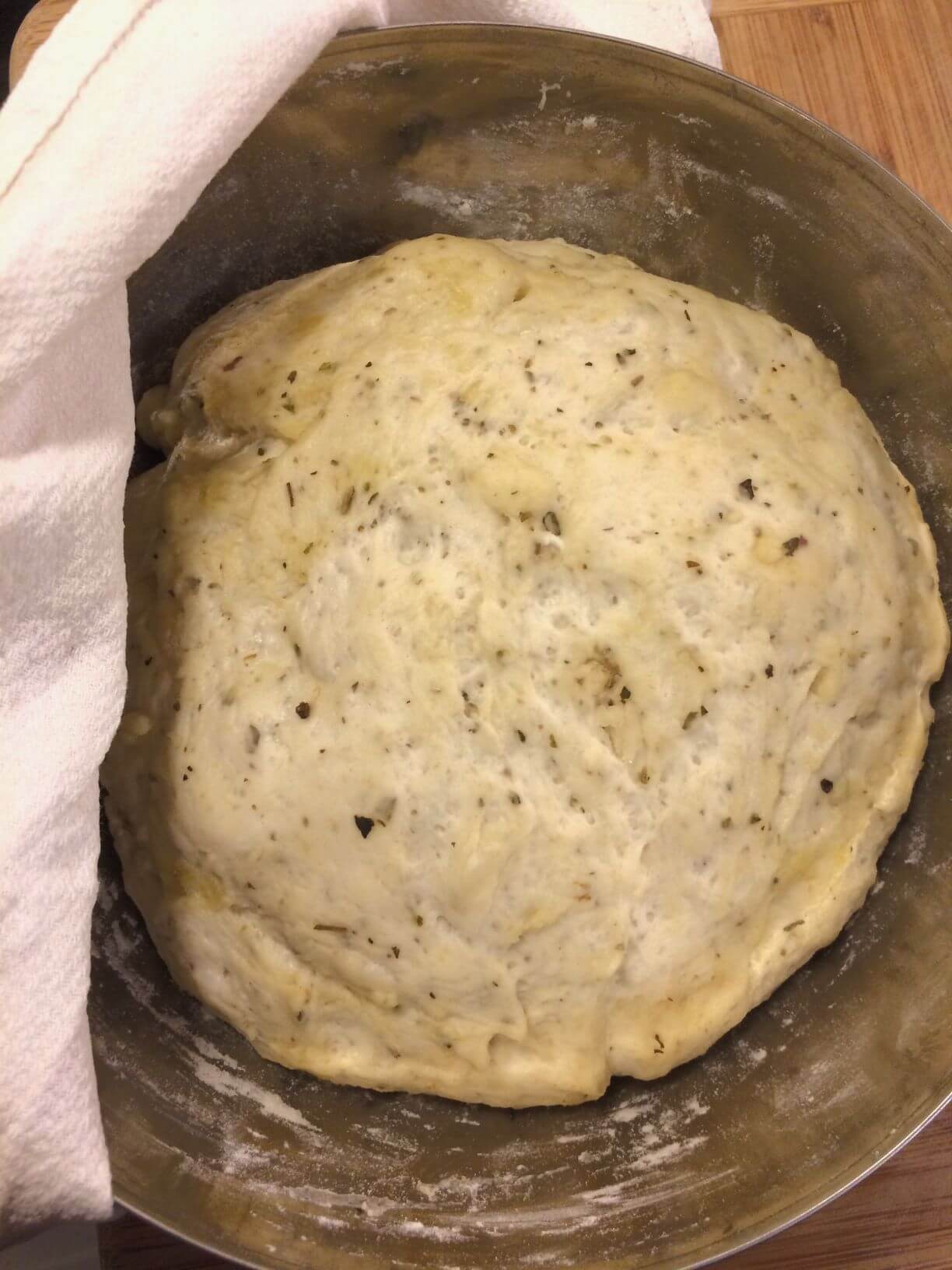 pizza dough with seasonings rising in a stainless steel bowl partially covered with tea towel, all on a wooden cutting board