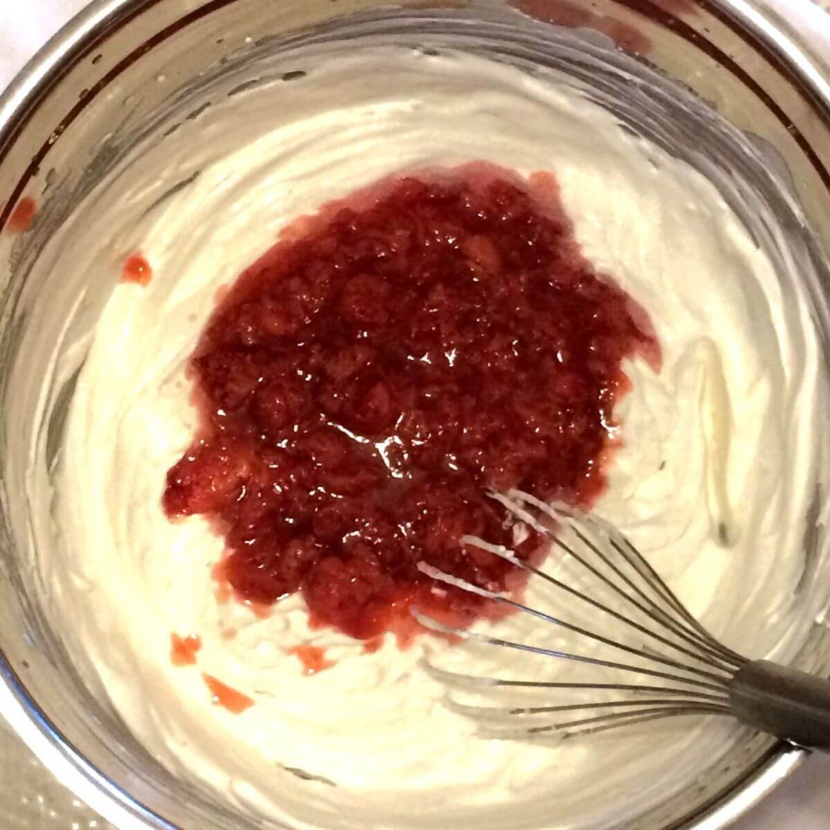 A large deep stainless steel bowl with whipped cream, coconut milk mix, and cooked strawberries added, with balloon whisk in bowl