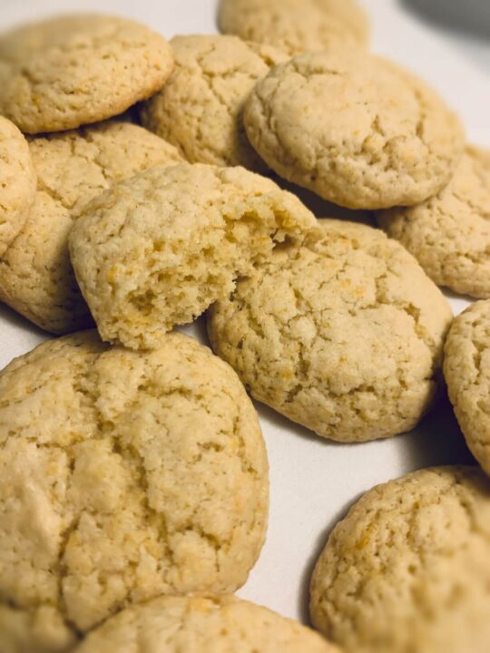 Baked lemon cookies with light golden yellow color and crackly tops piled on white plate with 1 cookie broken open to reveal chewy center