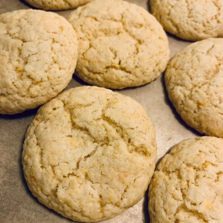 6 baked lemon cookies overlapping on parchment paper and cookie sheet with crackly top and light golden yellow color