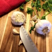 wooden cutting board with a full head of garlic, another head of garlic with the end cut off, garlic cloves with a knife laid on top of one, fresh cilantro leaves, and a red towel in the upper left corner