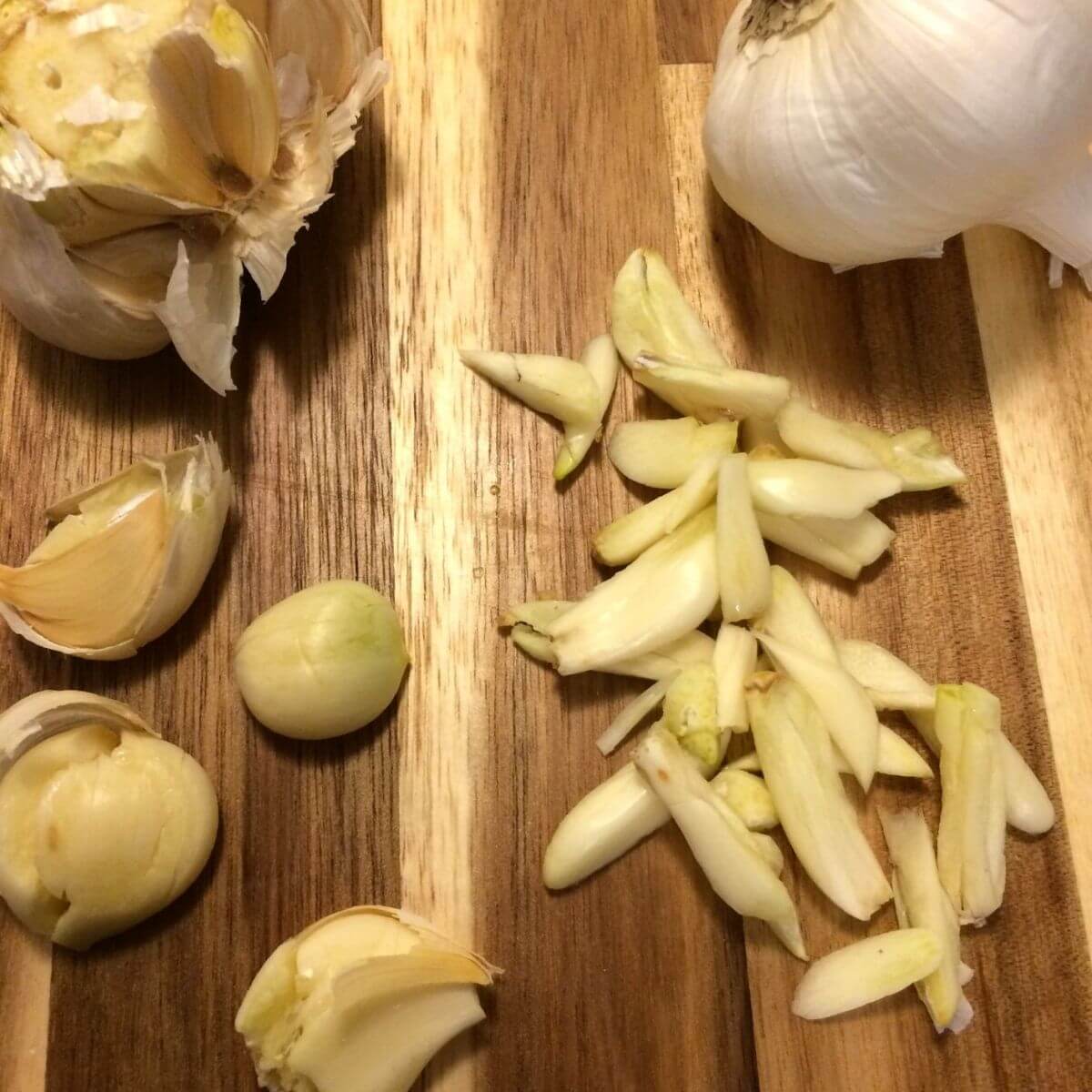 wooden cutting board with a full head of garlic, another head of garlic with the end cut off, 4 smashed garlic cloves semi-peeled, and sliced garlic cloves