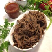 shredded beef piled on a white oval plate with fresh cilantro surrounding it, barbecue sauce in a small mason jar in the upper left corner and a red towel and Corning Ware bowl partially visible at the top of the image