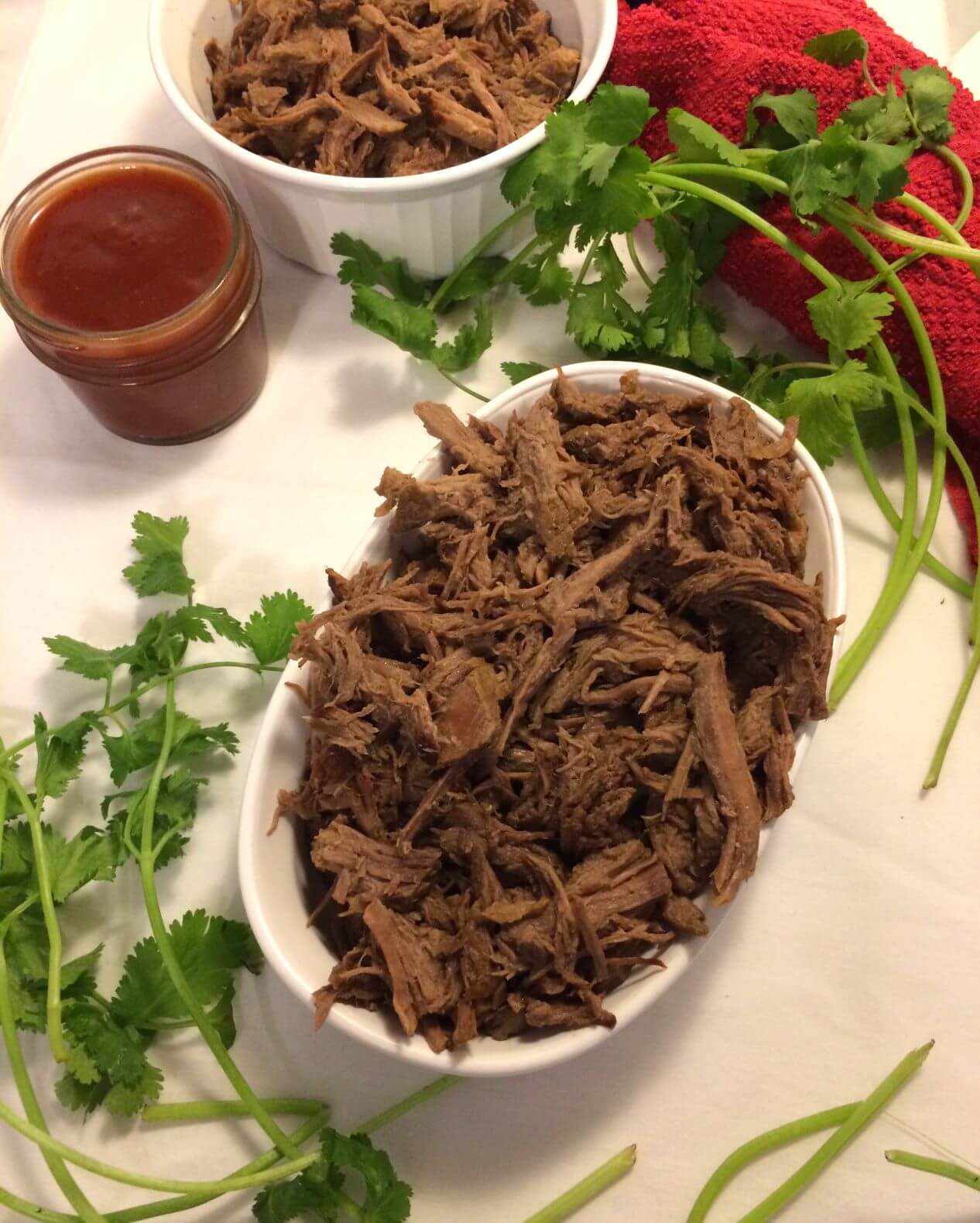shredded beef piled on a white oval plate with fresh cilantro surrounding it, barbecue sauce in a small mason jar in the upper left corner and a red towel and Corning Ware bowl with more shredded beef partially visible at the top of the image