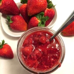 bird's eye view of jelly jar filled with strawberry jelly on white setting with an oval white plate with a pile of fresh large strawberries and a couple scattered strawberries on the table