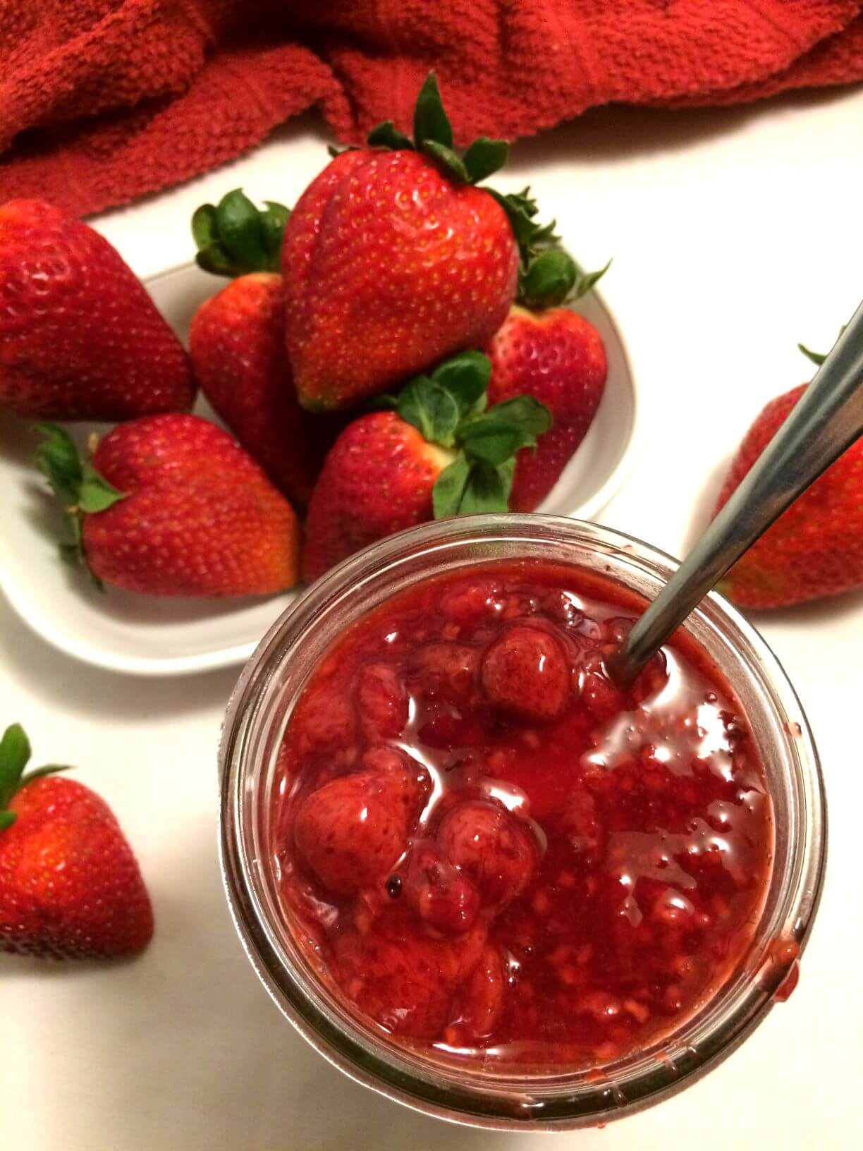 bird's eye view of jelly jar filled with strawberry jelly on white setting with an oval white plate with a pile of fresh large strawberries and a couple scattered strawberries on the table and a red towel in the top of the image