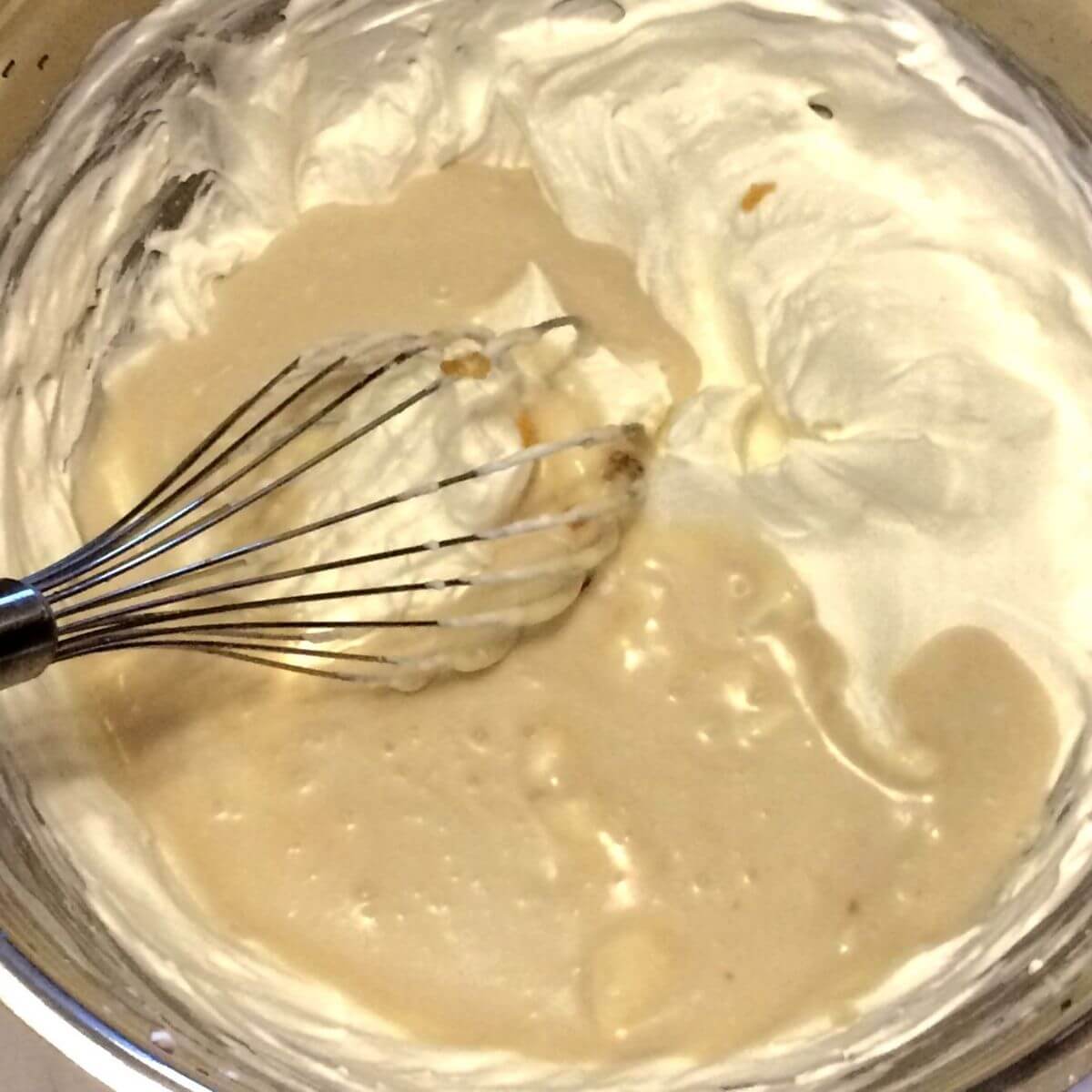 coconut milk and honey poured into whipped cream in large stainless steel bowl with balloon whisk sitting in bowl on left side