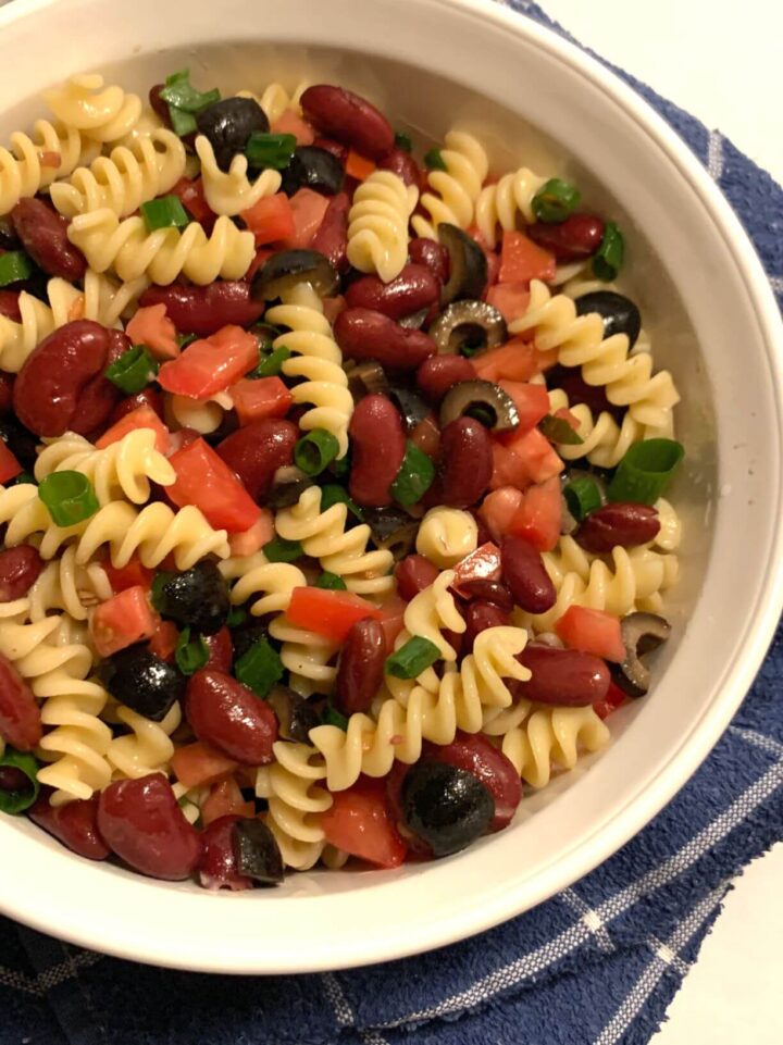 white Corning Ware bowl with fusilli noodles, diced black olives, dark red kidney beans, diced green onion, diced tomato mixed together, all on a blue and white towel.