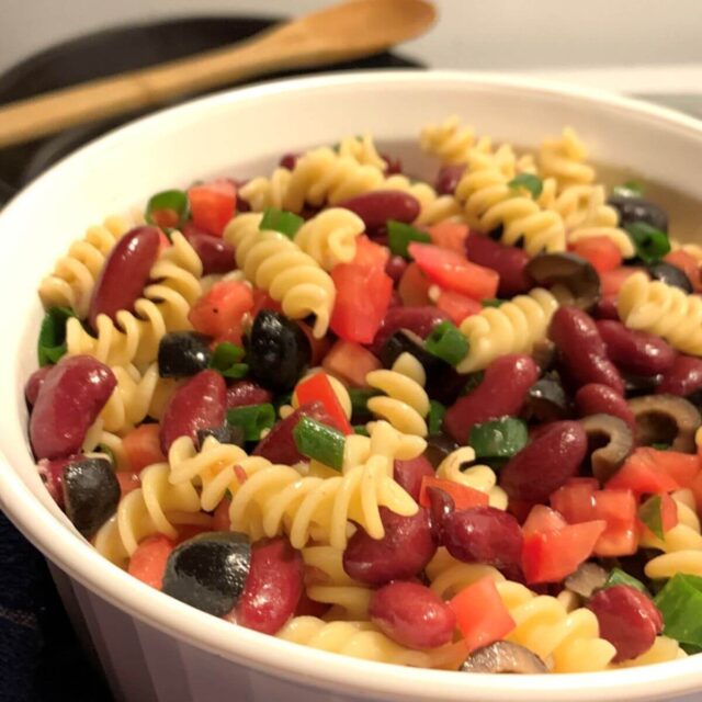 white Corning Ware bowl with fusilli noodles, diced black olives, dark red kidney beans, diced green onion, diced tomato mixed together. Wooden spoon in the background