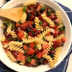 white Corning Ware bowl with fusilli noodles, diced black olives, dark red kidney beans, diced green onion, diced tomato mixed together with a wooden spoon in the bowl and all on a blue and white towel.