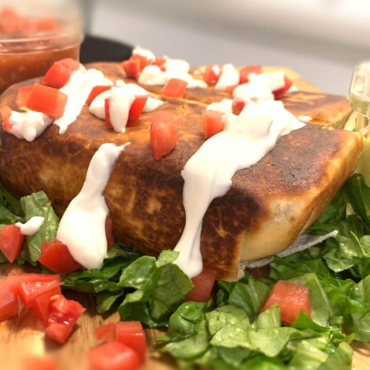 side view of a chimichanga with browned edges, topped with thinned Greek yogurt and diced tomatoes on a bed of chopped lettuce with jar of salsa in the back and a second chimichanga in the back.