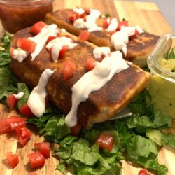 side view of a chimichanga with browned edges, topped with thinned Greek yogurt and diced tomatoes on a bed of chopped lettuce with jar of salsa in the upper left and guacamole on right side and a second chimichanga in the back all on a wooden cutting board