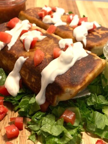 side view of a chimichanga with browned edges, topped with thinned Greek yogurt and diced tomatoes on a bed of chopped lettuce with jar of salsa in the upper left and guacamole on right side and a second chimichanga in the back all on a wooden cutting board