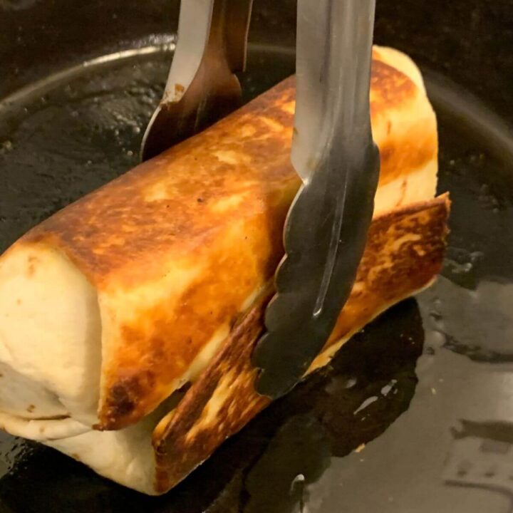 Chimichanga in cast iron skillet with olive oil held on edge with metal tongs.