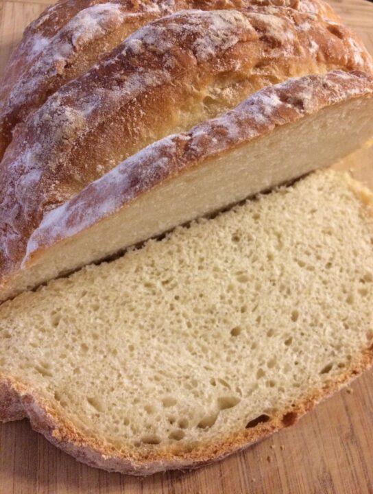 Close up of bloomer bread with deep slashes, flour dusting, lightly browned curst, cut open with a slice lying flat in front on cutting board.