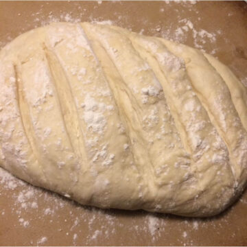 Elongated bloomer bread raw dough risen with deep slashes and flour dusting.