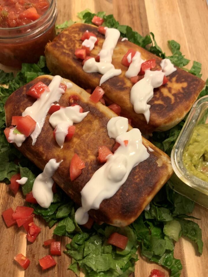 2 chimichangas with browned edges, topped with thinned Greek yogurt and diced tomatoes on a bed of chopped lettuce with jar of salsa in the upper left and guacamole on right side and a second chimichanga in the back all on a wooden cutting board