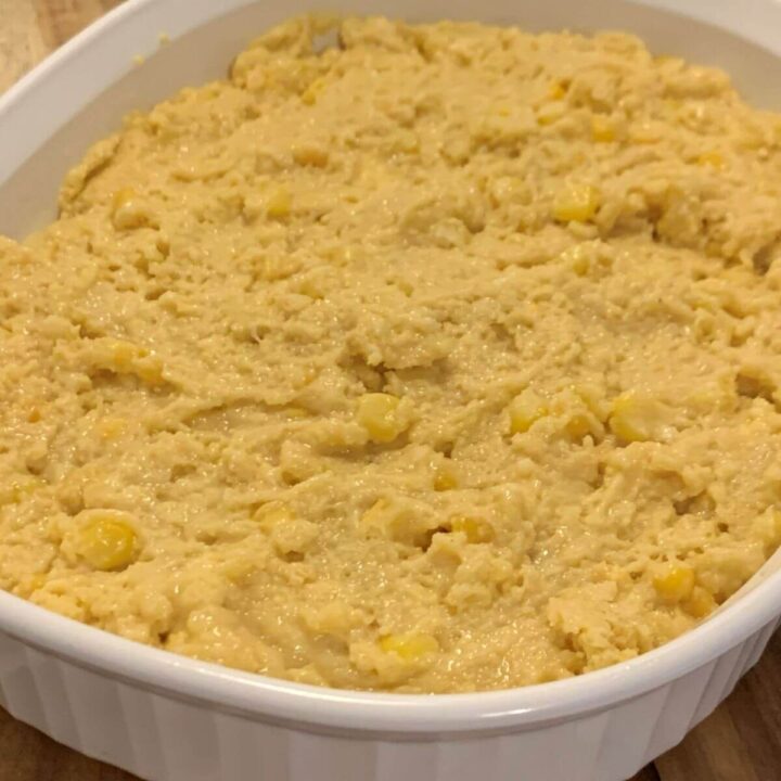 uncooked cornbread batter with corn kernels in a white CorningWare casserole dish all on a wooden cutting board.