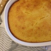 Square image of baked cornbread in a white CorningWare casserole dish with black and white striped towel in lower left corner and pie cutter partially in picture in upper left corner.