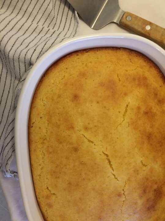 Vertical image of baked cornbread in white CorningWare casserole dish with white towel with black stripes in upper left corner and pie cutter in upper right corner.