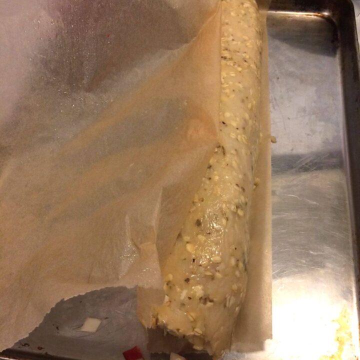 meat roll at edge of parchment paper, with parchment stuck on the left side of the roll, all on a stainless steel cookie sheet