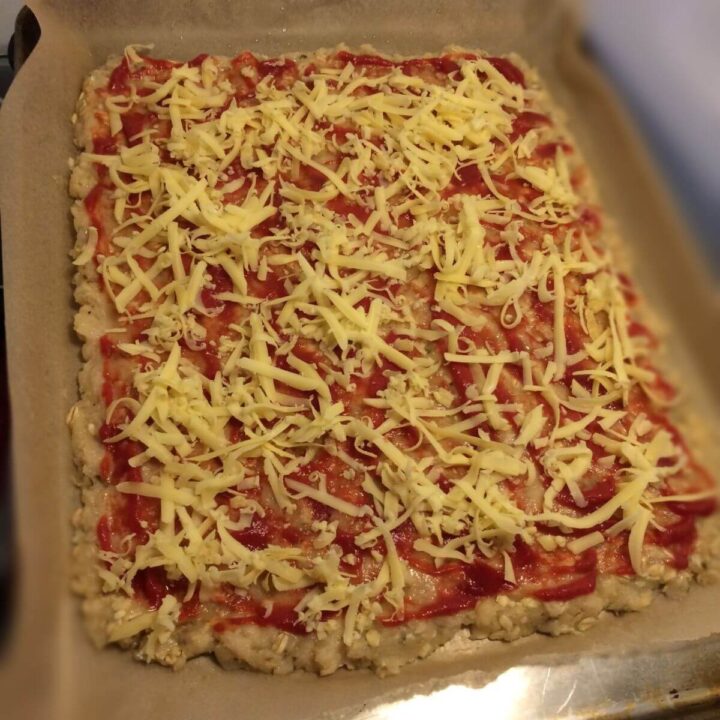 Ground meat pressed out flat on parchment paper with pizza sauce and shredded cheese spread over the top.