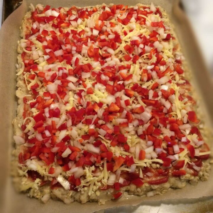 Ground meat pressed out flat on parchment paper with pizza sauce, shredded cheese, and diced vegetables spread over the top.