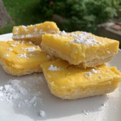 4 lemon bars cut into squares with powdered sugar dusted on top on a white plate outside with bushes blurred in the background