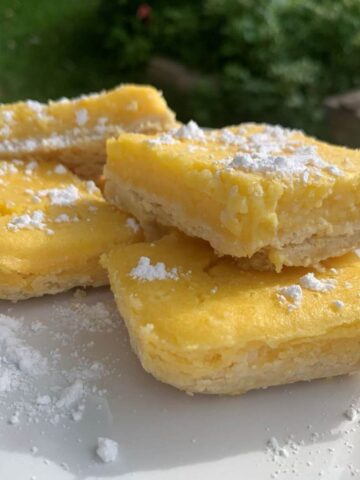 4 lemon bars cut into squares with powdered sugar dusted on top on a white plate outside with bushes blurred in the background